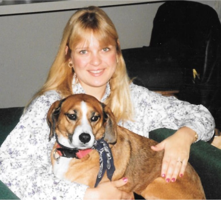 A woman with blonde hair sits on a couch, smiling at the camera, with her arm around a brown and white dog wearing a blue bandana.